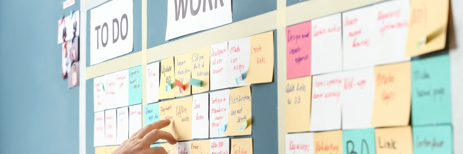 A green board covered with post-it notes. The notes are multiple colors, each with a task written on them. They are arranged, neatly, in a grid. There are two columns visible, to do and work. Someone points at one of the post-it notes.